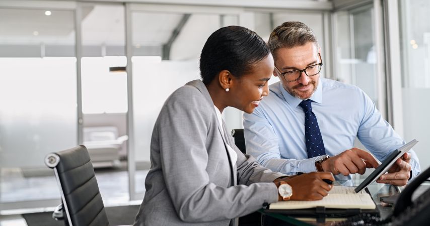 Homem e mulher, em uma mesa de escritório, analisando documentos na tela de um tablet e os dois com semblante de satisfação.