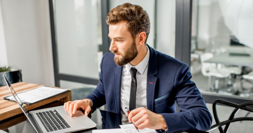 Homem de terno, analisando concentrado documentos em papel e no notebook, que está aberto em sua frente.
