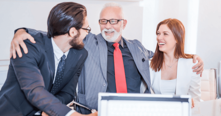 Homem de óculos, com barba e cabelos brancos, usando terno, sorrindo e abraçando um homem e uma mulher mais jovens.