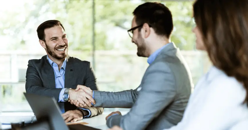 Imagem mostra dois homens brancos, usando terno, apertando as mãos em um ambiente empresarial. Os dois estão sorrindo. 
