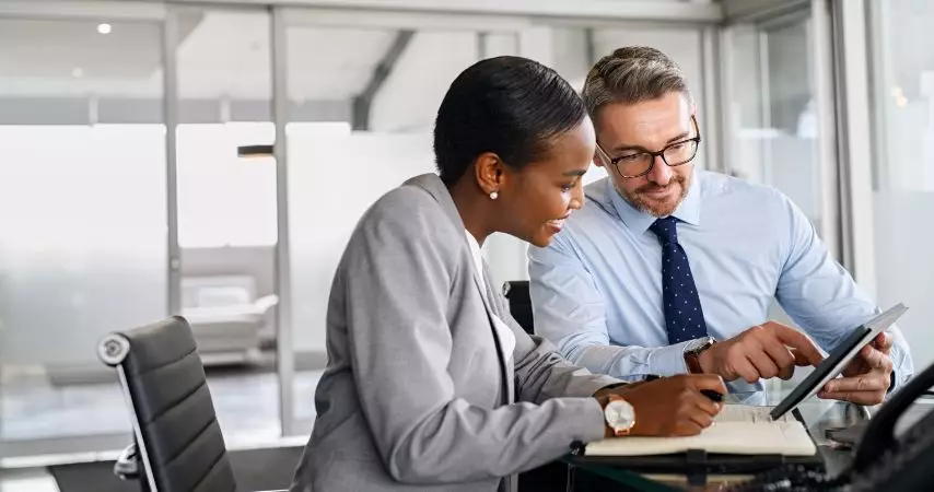 Homem e mulher, em uma mesa de escritório, analisando documentos na tela de um tablet e os dois com semblante de satisfação.