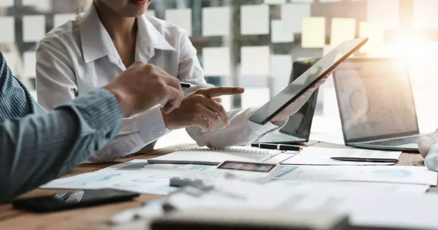 Homem e mulher em uma mesa de escritório cheia de documentos em cima. Eles analisam algo que está sendo mostrado na tela de um tablet.