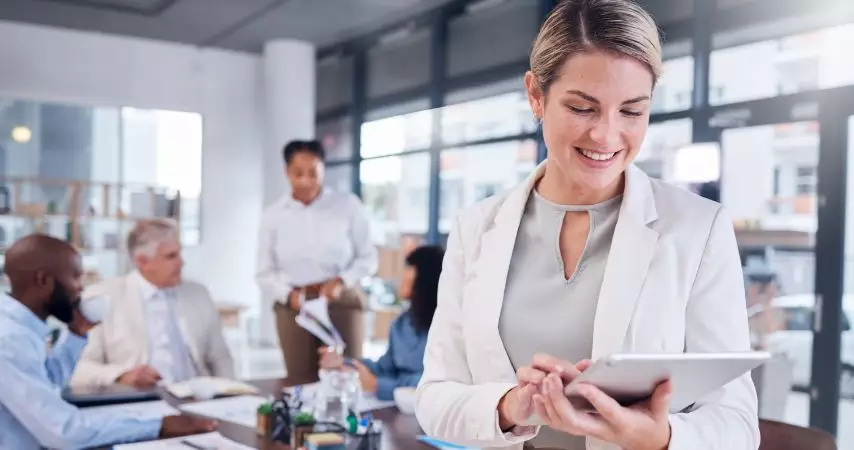 Em primeiro plano, mulher sorrindo e mexendo em um tablet. Ao fundo, equipe em reunião em uma mesa de escritório