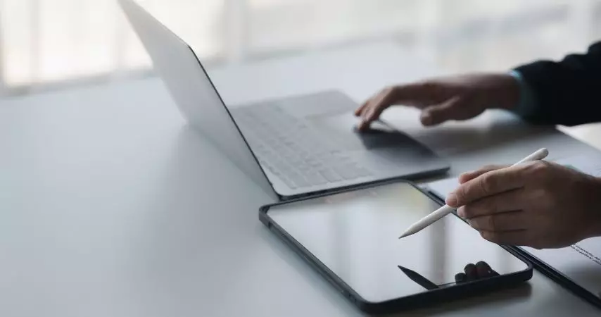 Homem mexendo em um tablet e em um notebook ao mesmo tempo, em um a mesa de escritório.
