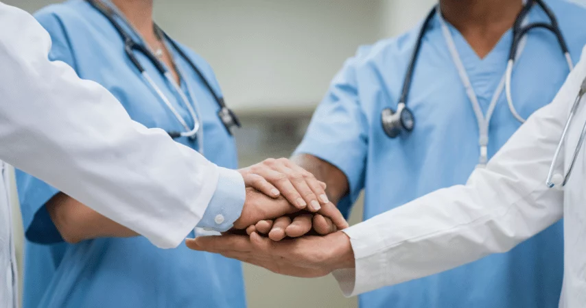 Quatro profisisonais de saúde com roupas de hospital colcoando a mão uma em cima da outra sinalizando união.