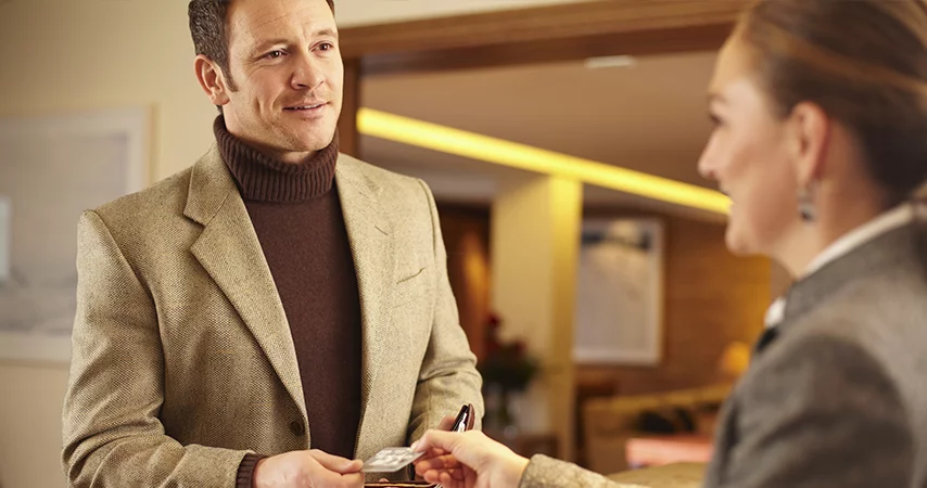 Homem de terno bege e blusão de gola rolê entregando cartão de crédito para recepcionista, mostrando a eficiencia de uma auditoria hoteleira.