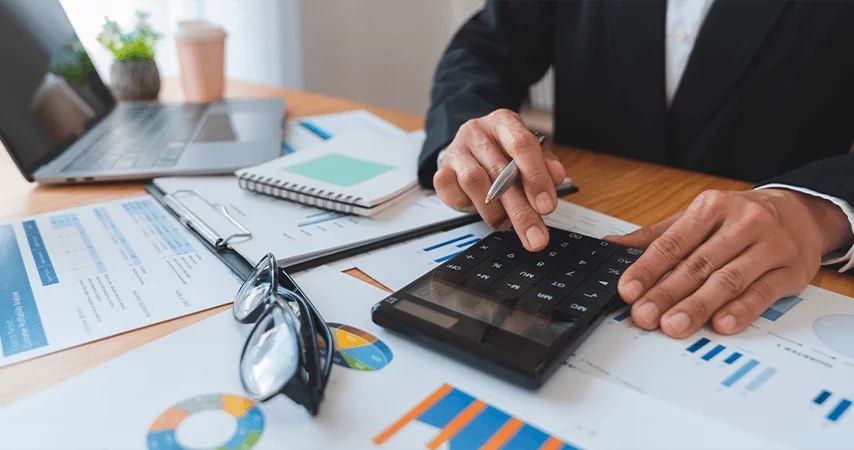 Imagem mostra homem de terno preto mexendo em uma calculadora em cima de uma mesa com muitos papéis com gráficos .