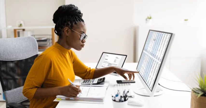 Mulher negra, vestindo uma blusa amarela, analisando dados em uma tela de computador, com um tablet ao lado e anotando em um documento. 