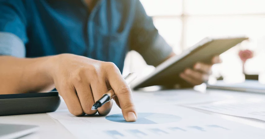 Homem com camisa social azul escuro com um tablet em uma das mãos e na outra uma caneta, apontando para um documento e calculadora ao lado.
