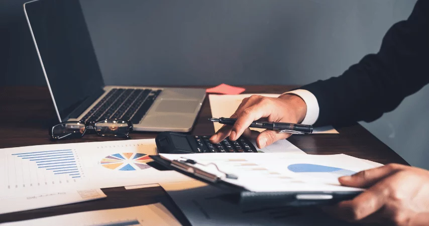 Imagem mostra os braços de um homem branco usando terno preto, com documentos contendo gráficos em diferentes formatos, usando uma calculadora e com uma caneta em uma das mãos. Na mesa também há um notebook com a tela aberta e um óculos ao lado do computador.