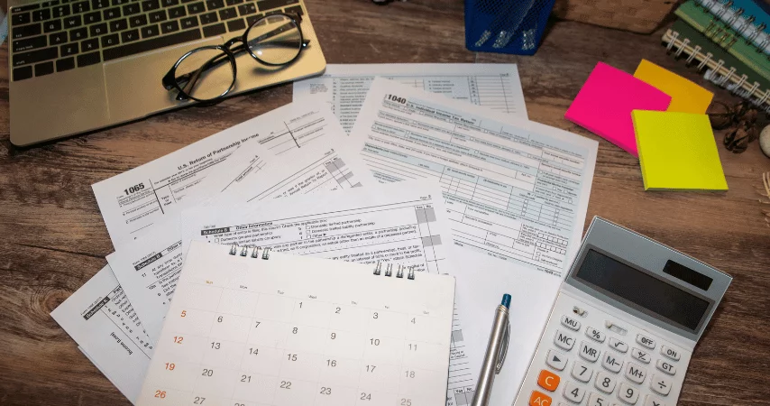 Documentos em folhas, calendário, calculadora, caneta, notebook aberto e óculos. Todos esses itens em cima da mesa.