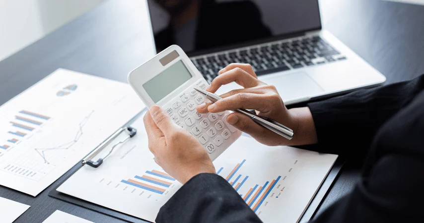 Homem de terno preto, utilizando uma calculadora, com gráficos em papéis em cima da mesa, junto com um notebook.